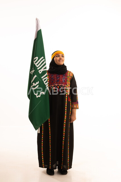 Anniversary of the founding of the first Saudi state, February 22, the day of our beginnings, 1727 AD, wearing the costumes of the central region, a portrait of a smiling Saudi Arabian Gulf woman wearing a kurta and a hat, holding the Founding Day flag in her hand, celebrating popular national occasions and holidays, full-length body, white background