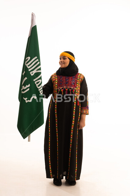 الاحتفال بالمناسبات والأعياد الوطنية الشعبية، ذكرى تأسيس الدولة السعودية الأولى 22 فبراير، يوم بدينا 1727 م، ارتداء أزياء المنطقة الوسطى، بورتريه لإمرأة عربية خليجية سعودية مبتسمة ترتدي الكرتة والهامة تحمل بيدها علم يوم التأسيس، الجسم بالطول الكامل، خلفية بيضاء