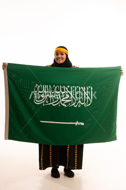 الفخر بالتراث التاريخي، يوم العلم 11 مارس، يوم بدينا 1727م, ارتداء زي المنطقة الوسطى، اليوم الوطني السعودي 23 سبتمبر, بورتريه لإمرأة عربية خليجية سعودية ترتدي الكرتة والهامة تحمل علم المملكة تنظر إلى الكاميرا بتعابير الفخر, الجسم بالطول الكامل، خلفية بيضاء