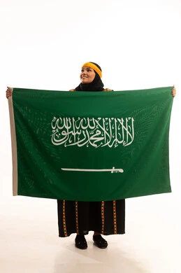 الاعتزاز والتباهي بإنجازات البلاد، الفخر بالتراث التاريخي، يوم العلم 11 مارس، يوم بدينا 1727م, ارتداء زي المنطقة الوسطى، اليوم الوطني السعودي 23 سبتمبر, بورتريه لإمرأة عربية خليجية سعودية ترتدي الكرتة والهامة تحمل علم المملكة, الجسم بالطول الكامل، خلفية بيضاء