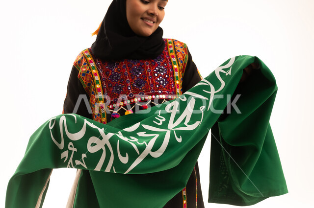 الاحتفال بالمناسبات الوطنية، مفهوم حب الوطن، اليوم الوطني السعودي 23 سبتمبر, يوم بدينا 1727م, الفخر بالتراث التاريخي، يوم العلم 11 مارس، ارتداء زي المنطقة الوسطى، بورتريه مقرب لإمرأة عربية خليجية سعودية ترتدي الكرتة والهامة تحمل علم المملكة, خلفية بيضاء