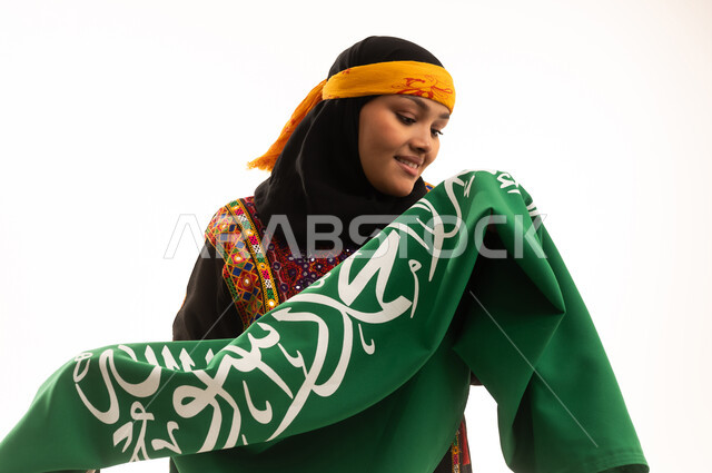 Celebrating national occasions, the concept of patriotism, Saudi National Day September 23, the day we started in 1727 AD, pride in historical heritage, Flag Day March 11, wearing the Central Region costume, close-up portrait of a Saudi Arabian Gulf woman wearing a kurta and a hat holding the Kingdom's flag, white background