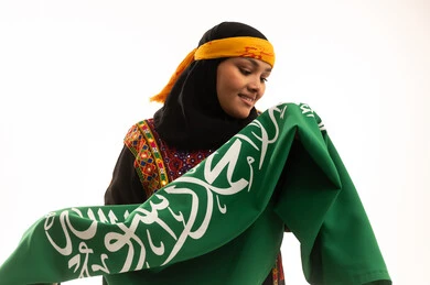 الاحتفال بالمناسبات الوطنية، مفهوم حب الوطن، اليوم الوطني السعودي 23 سبتمبر, يوم بدينا 1727م, الفخر بالتراث التاريخي، يوم العلم 11 مارس، ارتداء زي المنطقة الوسطى، بورتريه مقرب لإمرأة عربية خليجية سعودية ترتدي الكرتة والهامة تحمل علم المملكة, خلفية بيضاء