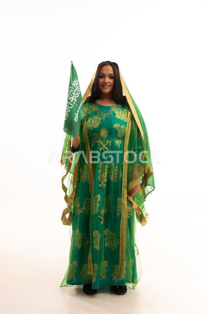 Looking at the camera with gestures of pride, Flag Day March 11, Badina Day February 22, wearing the traditional costume of the central region on a national occasion, Saudi National Day September 23, portrait of a Saudi Arabian Gulf woman wearing a sheila and bisht holding the flag of the Kingdom, full body length, white background