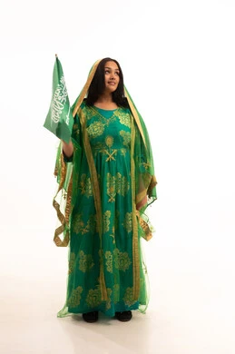 الوقوف باستقامة بإيماءات الشموخ، ارتداء الزي الشعبي للمنطقة الوسطى في مناسبة وطنية, يوم العلم 11 مارس، يوم بدينا22 فبراير, اليوم الوطني السعودي 23 سبتمبر، بورتريه لامرأة عربية خليجية سعودية ترتدي الشيلة والبشت تمسك علم المملكة، الطول الكامل للجسم، خلفية بيضاء