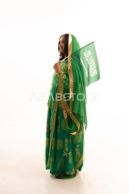 اليوم الوطني السعودي 23 سبتمبر، رفع راية التوحيد والإسلام، يوم العلم 11 مارس، ارتداء الزي الشعبي للمنطقة الوسطى في مناسبة وطنية, يوم بدينا22 فبراير, بورتريه من الجانب لامرأة عربية خليجية سعودية ترتدي الشيلة والبشت تمسك علم المملكة، الطول الكامل للجسم، خلفية بيضاء