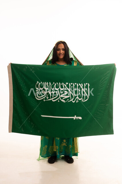 النظر إلى الكاميرا بإيماءات الاعتزاز، يوم العلم 11 مارس، يوم بدينا22 فبراير, ارتداء الزي الشعبي للمنطقة الوسطى في مناسبة وطنية, اليوم الوطني السعودي 23 سبتمبر، بورتريه لامرأة عربية خليجية سعودية ترتدي الشيلة والبشت تمسك علم المملكة، الطول الكامل للجسم، خلفية بيضاء