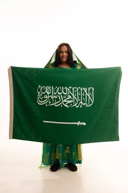 النظر إلى الكاميرا بإيماءات الاعتزاز، يوم العلم 11 مارس، يوم بدينا22 فبراير, ارتداء الزي الشعبي للمنطقة الوسطى في مناسبة وطنية, اليوم الوطني السعودي 23 سبتمبر، بورتريه لامرأة عربية خليجية سعودية ترتدي الشيلة والبشت تمسك علم المملكة، الطول الكامل للجسم، خلفية بيضاء