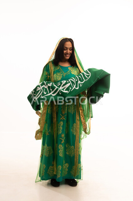 Standing upright with proud gestures, wearing the traditional costume of the central region on a national occasion, Flag Day March 11, Badina Day February 22, Saudi National Day September 23, portrait of a Saudi Arabian Gulf woman wearing a sheila and bisht holding the flag of the Kingdom, full body length, white background