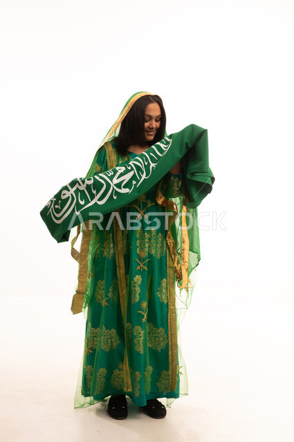 Flag Day, March 11, standing upright with proud gestures, wearing the traditional costume of the central region on a national occasion, Badina Day, February 22, Saudi National Day, September 23, portrait of a Saudi Arabian Gulf woman wearing a sheila and bisht holding the flag of the Kingdom, full body length, white background