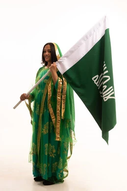 الوقوف باستقامة ورفع علم التأسيس بتعابير الشموخ والتباهي، ايماءات البهجة والسرور، ذكرى تأسيس الدولة السعودية الاولى ٢٢ فبراير، بورتريه لامرأة عربية خليجية سعودية ترتدي الشيلة والبشت، الاحتفال بيوم بدينا ١٧٢٧م، صورة لكامل الجسم، خلفية بيضاء