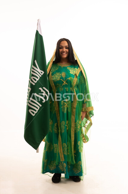 النظر الى الكاميرا بايماءات البهجة والسرور، ذكرى تأسيس الدولة السعودية الاولى ٢٢ فبراير، بورتريه لامرأة عربية خليجية سعودية ترتدي الشيلة والبشت، الاحتفال بيوم بدينا ١٧٢٧م، رفع علم التأسيس بتعابير الشموخ والتباهي، صورة لكامل الجسم، خلفية بيضاء