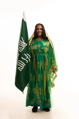 النظر الى الكاميرا بايماءات البهجة والسرور، ذكرى تأسيس الدولة السعودية الاولى ٢٢ فبراير، بورتريه لامرأة عربية خليجية سعودية ترتدي الشيلة والبشت، الاحتفال بيوم بدينا ١٧٢٧م، رفع علم التأسيس بتعابير الشموخ والتباهي، صورة لكامل الجسم، خلفية بيضاء