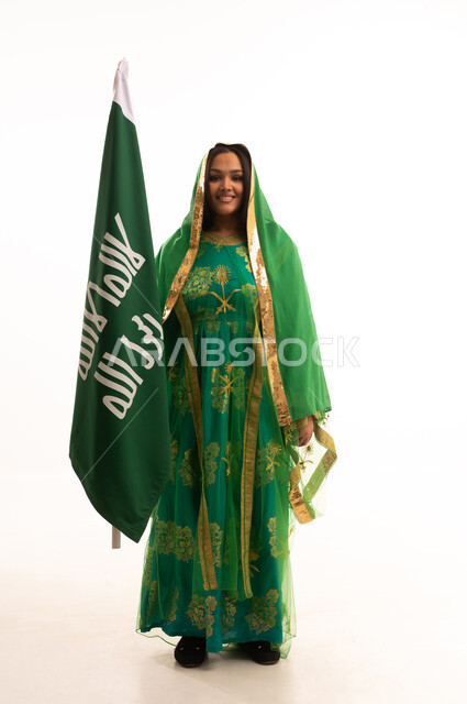 النظر الى الكاميرا بايماءات البهجة والسرور، ذكرى تأسيس الدولة السعودية الاولى ٢٢ فبراير، بورتريه لامرأة عربية خليجية سعودية ترتدي الشيلة والبشت، الاحتفال بيوم بدينا ١٧٢٧م، رفع علم التأسيس بتعابير الشموخ والتباهي، صورة لكامل الجسم، خلفية بيضاء