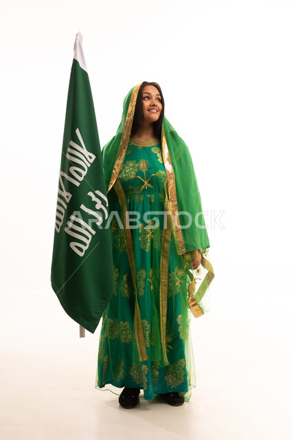 Celebrating the day of our beginnings in 1727 AD, standing upright and raising the founding flag with expressions of pride and boasting, gestures of joy and happiness, the anniversary of the founding of the first Saudi state, February 22, portrait of a Saudi Arabian Gulf woman wearing a sheila and bisht, full body image, white background