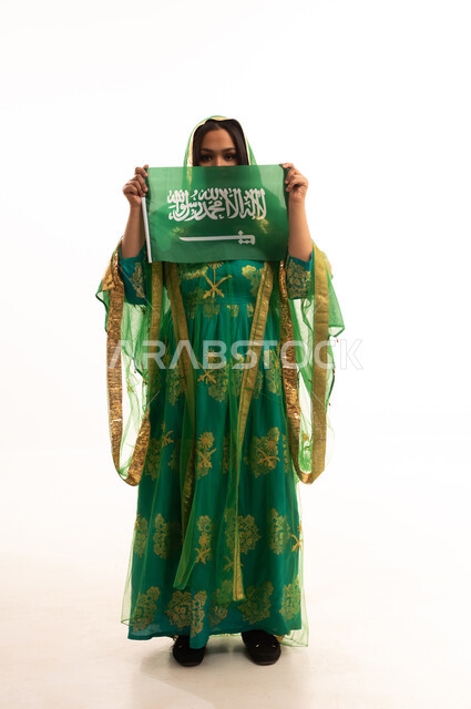 النظر إلى الكاميرا بإيماءات الاعتزاز، يوم العلم 11 مارس، يوم بدينا22 فبراير, ارتداء الزي الشعبي للمنطقة الوسطى في مناسبة وطنية, اليوم الوطني السعودي 23 سبتمبر، بورتريه لامرأة عربية خليجية سعودية ترتدي الشيلة والبشت تمسك علم المملكة، الطول الكامل للجسم، خلفية بيضاء