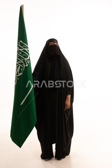التطلع والنظر إلى الأعلى بتعابير الشموخ والفخر، اليوم الوطني السعودي 23 سبتمبر، ارتداء الزي الشعبي في مناسبة وطنية، يوم العلم 11 مارس، يوم بدينا22 فبراير, بورتريه لامرأة عربية خليجية سعودية ترتدي البرقع والعباءة تمسك علم المملكة، الطول الكامل للجسم، خلفية بيضاء