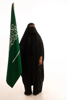 التطلع والنظر إلى الأعلى بتعابير الشموخ والفخر، اليوم الوطني السعودي 23 سبتمبر، ارتداء الزي الشعبي في مناسبة وطنية، يوم العلم 11 مارس، يوم بدينا22 فبراير, بورتريه لامرأة عربية خليجية سعودية ترتدي البرقع والعباءة تمسك علم المملكة، الطول الكامل للجسم، خلفية بيضاء