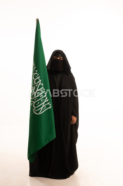 رفع راية التوحيد والإسلام، يوم العلم 11 مارس، يوم بدينا22 فبراير, ارتداء الزي الشعبي في مناسبة وطنية, الاحتفال بذكرى اليوم الوطني السعودي 23 سبتمبر، بورتريه لامرأة عربية خليجية سعودية ترتدي البرقع والعباءة تمسك علم المملكة، الطول الكامل للجسم، خلفية بيضاء