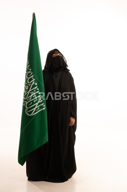التطلع والنظر إلى الأعلى بتعابير الشموخ والفخر، اليوم الوطني السعودي 23 سبتمبر، ارتداء الزي الشعبي في مناسبة وطنية، يوم العلم 11 مارس، يوم بدينا22 فبراير, بورتريه لامرأة عربية خليجية سعودية ترتدي البرقع والعباءة تمسك علم المملكة، الطول الكامل للجسم، خلفية بيضاء