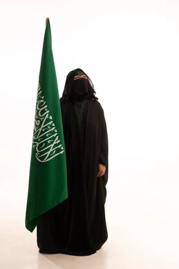التطلع والنظر إلى الأعلى بتعابير الشموخ والفخر، اليوم الوطني السعودي 23 سبتمبر، ارتداء الزي الشعبي في مناسبة وطنية، يوم العلم 11 مارس، يوم بدينا22 فبراير, بورتريه لامرأة عربية خليجية سعودية ترتدي البرقع والعباءة تمسك علم المملكة، الطول الكامل للجسم، خلفية بيضاء