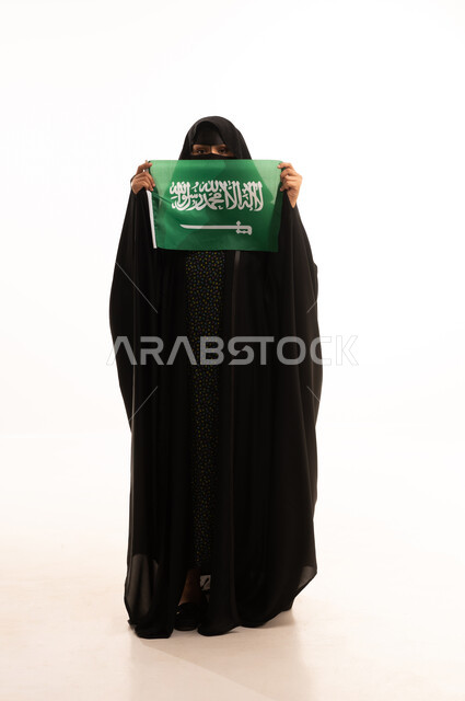 الاحتفال بذكرى اليوم الوطني السعودي 23 سبتمبر، ارتداء الزي الشعبي في مناسبة وطنية، يوم العلم 11 مارس، يوم بدينا22 فبراير, بورتريه لامرأة عربية خليجية سعودية ترتدي البرقع والعباءة تمسك علم المملكة وتنظر إلى الكامير بتعابير الاعتزاز، الطول الكامل للجسم، خلفية بيضاء