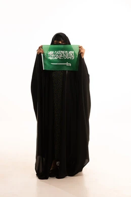 الاحتفال بذكرى اليوم الوطني السعودي 23 سبتمبر، ارتداء الزي الشعبي في مناسبة وطنية، يوم العلم 11 مارس، يوم بدينا22 فبراير, بورتريه لامرأة عربية خليجية سعودية ترتدي البرقع والعباءة تمسك علم المملكة وتنظر إلى الكامير بتعابير الاعتزاز، الطول الكامل للجسم، خلفية بيضاء