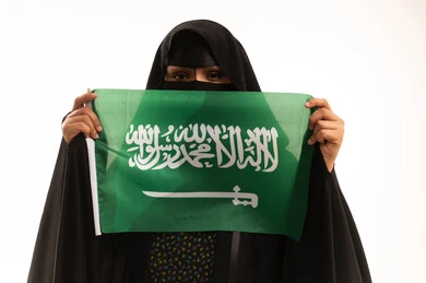 إظهاء الولاء للبلاد، يوم بدينا22 فبراير, اليوم الوطني السعودي 23 سبتمبر، ارتداء الزي الشعبي في مناسبة وطنية، يوم العلم 11 مارس، بورتريه مقرب لامرأة عربية خليجية سعودية ترتدي البرقع والعباءة تمسك علم المملكة وتنظر إلى الكاميرا بإيماءات الثقة بالنفس، خلفية بيضاء