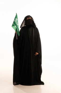 Looking up with expressions of pride and dignity, Saudi National Day September 23, wearing traditional dress on a national occasion, Flag Day March 11, Badina Day February 22, portrait of a Saudi Arabian Gulf woman wearing a burqa and abaya holding the flag of the Kingdom, full body length, white background