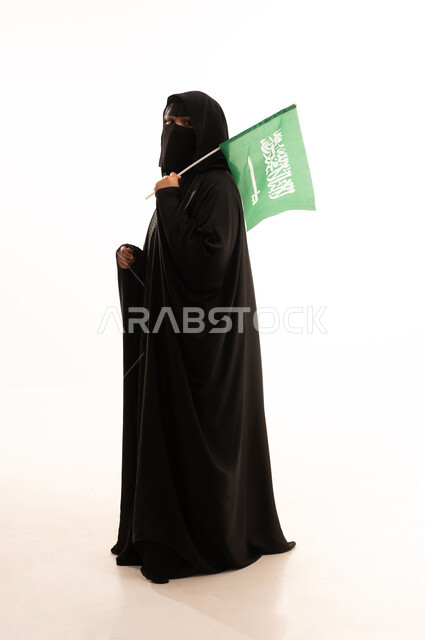 يوم بدينا22 فبراير, النظر إلى الكاميرا بتعابير الاعتزاز، اليوم الوطني السعودي 23 سبتمبر، ارتداء الزي الشعبي في مناسبة وطنية، يوم العلم 11 مارس، بورتريه من الجانب لامرأة عربية خليجية سعودية ترتدي البرقع والعباءة تمسك علم المملكة، الطول الكامل للجسم، خلفية بيضاء