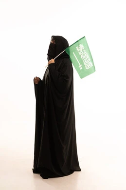 Looking up with expressions of pride and dignity, Saudi National Day September 23, wearing traditional dress on a national occasion, Flag Day March 11, Badina Day February 22, side portrait of a Saudi Arabian Gulf woman wearing a burqa and abaya holding the Kingdom's flag, full body length, white background