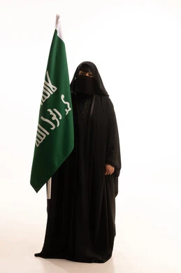 إظهار الولاء والحب للوطن, ذكرى تأسيس الدولة السعودية الأولى 1727م، بورتريه لامرأة عربية خليجية سعودية ترتدي البرقع والعباءة تحمل علم التأسيس تنظر إلى الكاميرا بإيماءات الاعتزاز والشموخ، الفخر بإنجازات المملكة، يوم بدينا 22 فبراير، الجسم بالطول الكامل، خلفية بيضاء