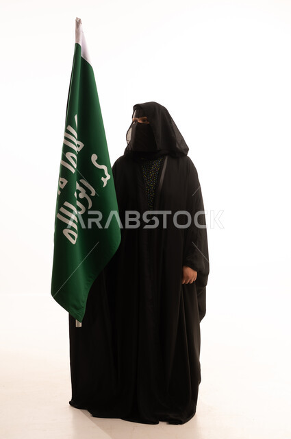 ذكرى تأسيس الدولة السعودية الأولى 1727م، الفخر بإنجازات المملكة، إظهار الولاء والحب للوطن, بورتريه لامرأة عربية خليجية سعودية ترتدي البرقع والعباءة تحمل علم التأسيس بإيماءات الاعتزاز والشموخ، يوم بدينا 22 فبراير، الجسم بالطول الكامل، خلفية بيضاء