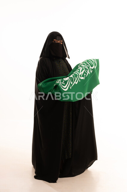 Raising the flag of unity and Islam, Flag Day March 11, Badina Day February 22, wearing traditional dress on a national occasion, celebrating Saudi National Day September 23, portrait of a Saudi Arabian Gulf woman wearing a burqa and abaya holding the flag of the Kingdom, full body length, white background