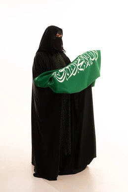 Pride and pride in belonging to the country, our beginning day, February 22, Saudi National Day, September 23, wearing traditional dress on a national occasion, Flag Day, March 11, portrait of a Saudi Arabian Gulf woman wearing a burqa and abaya holding the flag of the Kingdom, full body length, white background