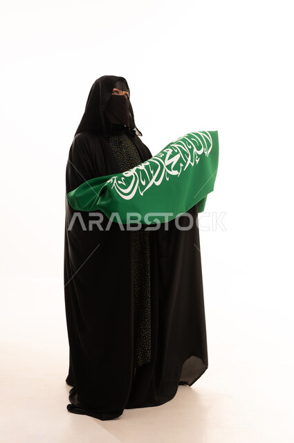 التطلع والنظر إلى الأعلى بتعابير الشموخ والفخر، اليوم الوطني السعودي 23 سبتمبر، ارتداء الزي الشعبي في مناسبة وطنية، يوم العلم 11 مارس، يوم بدينا22 فبراير, بورتريه لامرأة عربية خليجية سعودية ترتدي البرقع والعباءة تمسك علم المملكة، الطول الكامل للجسم، خلفية بيضاء