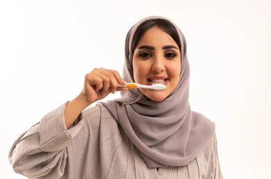 ابتسامة ناصعة البياض، الحفاظ على الأسنان وتنظيفها بشكل مستمر, مفهوم العادات الصحية, روتين نظافة الفم اليومية، تجنب التسوس وأمراض اللثة, بورتريه مقرب لامرأة عربية خليجية سعودية ترتدي الحجاب و العباءة تقوم بغسل اسنانها باستخدام الفرشاة ، خلفية بيضاء