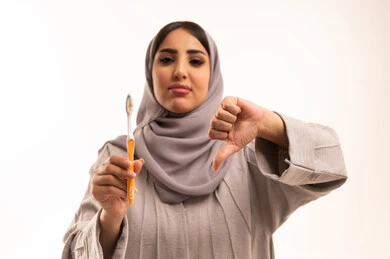 الإشارة بعلامة عدم القبول والجودة السيئة، روتين نظافة الفم اليومية، مفهوم العناية والاهتمام بصحة ونظافة الأسنان، بورتريه مقرب لامرأة عربية خليجية سعودية ترتدي الحجاب و العباءة تقوم بيديها فرشاة اسنان، خلفية بيضاء