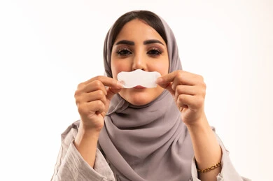 Getting rid of blackheads and purifying the skin, caring for and improving skin freshness, using pore-cleansing masks and cosmetic ingredients, a close-up portrait of a Saudi Gulf Arab woman wearing a hijab and abaya applying nose strips, white background.