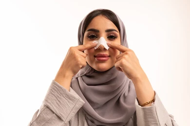 Getting rid of blackheads and dead skin, a close-up portrait of a Saudi Arabian Gulf woman wearing a hijab and abaya applying nose strips, using pore cleansing masks and cosmetic ingredients, taking care of and focusing on skin freshness and improvement, white background.