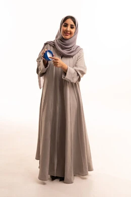 النظر للكاميرا بتعابير السرور، تحسين مظهر الجلد من خلال الترطيب والتنظيف، استخدام مكونات تجميلية شبابية، بورتريه لامرأة عربية خليجية سعودية ترتدي الحجاب و العباءة تضع كريم المرطب على يديها، الجسم بالطول الكامل، خلفية بيضاء