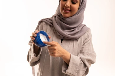 Skincare through moisturizing, cleansing, and brightening, using youthful cosmetic ingredients, a close-up portrait of a Saudi Arabian Gulf woman wearing a hijab and abaya applying moisturizer, white background.