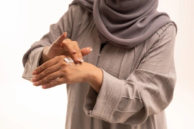 ترطيب وتنظيف وتفتيح الجلد، روتين العناية بالبشرة ، مفهوم الإهتمام بنضارة وتحسين البشرة، استخدام مكونات تجميلية شبابية، بورتريه مقرب لامرأة عربية خليجية سعودية ترتدي الحجاب و العباءة تضع كريم المرطب على يديها، خلفية بيضاء