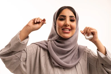 تجنب التسوس وأمراض اللثة، روتين نظافة الفم اليومية، التأكد من نظافة الأسنان بعد تناول الطعام, بورتريه مقرب لامرأة عربية خليجية سعودية ترتدي الحجاب و العباءة تقوم باستعمال الخيط السني، النظر الى الكاميرا بإيماءات السعادة والسرور، خلفية بيضاء