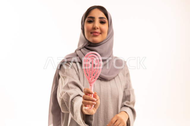 ترتيب وتمشيط الشعر، المحافظة على شعر صحي وأنيق، مفهوم العناية والاهتمام بالجمال والأناقة والمظهر الخارجي، بورتريه مقرب لامرأة عربية خليجية سعودية ترتدي الحجاب و العباءة تمسك بيدها فرشاة الشعر ( المشط ) بتعابير وإيماءات تدل على السعادة والسرور، خلفية بيضاء