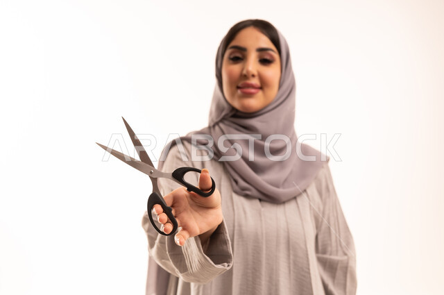 قص و تصفيف الشعر في صالون الحلاقة، النظر إلى الكاميرا بإيماءات السرور، مركز تجميلي نسائي، مفهوم العناية و الاهتمام بالجمال والمظهر الخارجي، بورتريه مقرب لامرأة عربية خليجية سعودية ترتدي الحجاب و العباءة تحمل بيدها المقص، خلفية بيضاء