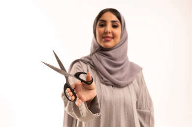 قص و تصفيف الشعر في صالون الحلاقة، النظر إلى الكاميرا بإيماءات السرور، مركز تجميلي نسائي، مفهوم العناية و الاهتمام بالجمال والمظهر الخارجي، بورتريه مقرب لامرأة عربية خليجية سعودية ترتدي الحجاب و العباءة تحمل بيدها المقص، خلفية بيضاء