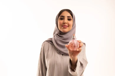 التزيين والتجمل النسائي, بورتريه مقرب لامرأة عربية خليجية سعودية  ترتدي الحجاب و العباءة تحمل بيدها زجاجة عطر عصرية, إيماءات السعادة لشراء زيوت عطرية جديدة, التسويق لمنتجات سعودية وعالمية, خلفية بيضاء