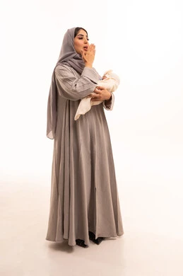 The need for sleep and rest, feelings of compassion and tenderness, strengthening the bond of motherhood, caring for and nurturing children, a portrait of an Arab Gulf Saudi woman wearing an abaya and hijab holding her daughter and yawning, full-body, white background.