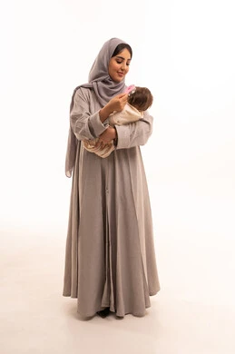 مفهوم الأمومة والطفولة، حنان الأمهات و تحمل المسؤولية والعناية بالأطفال، بورترية لامراه عربيه خليجيه سعودية ترتدي العباءة و الحجاب تحمل طفلتها الرضيعة و تقوم بتمشيط شعرها باستخدام المشط، الجسم بالطول الكامل، خلفية بيضاء